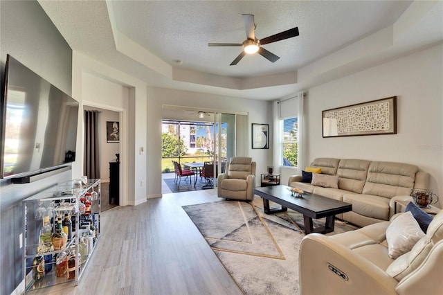 living area with a raised ceiling, a textured ceiling, wood finished floors, baseboards, and ceiling fan