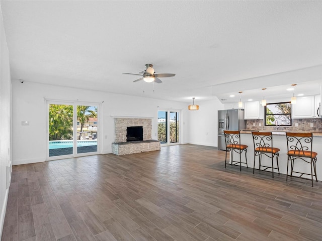 unfurnished living room featuring a sink, dark wood-style floors, a stone fireplace, baseboards, and ceiling fan