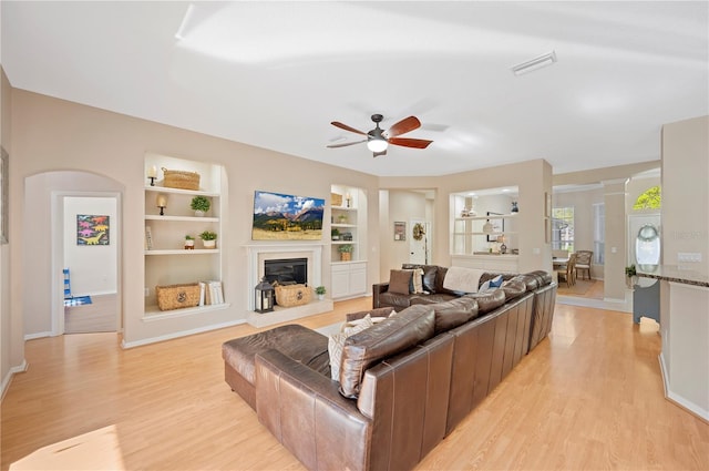 living area featuring built in features, a glass covered fireplace, a ceiling fan, and light wood finished floors