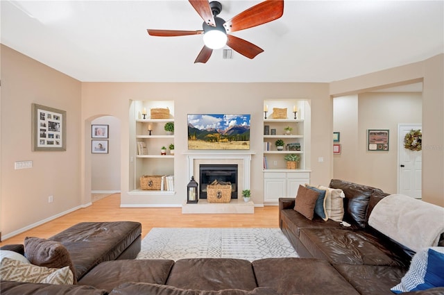 living room featuring a glass covered fireplace, built in shelves, light wood-style floors, and a ceiling fan