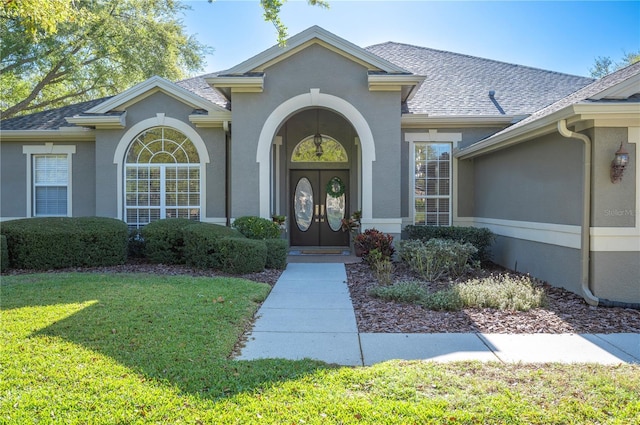 entrance to property featuring a shingled roof, a yard, and stucco siding