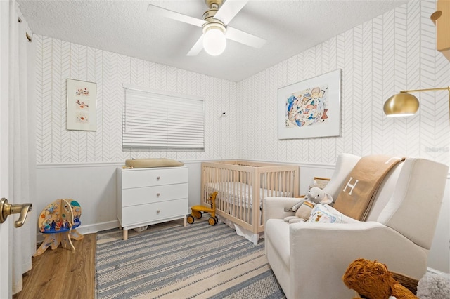 bedroom featuring a crib, wallpapered walls, ceiling fan, wood finished floors, and a textured ceiling