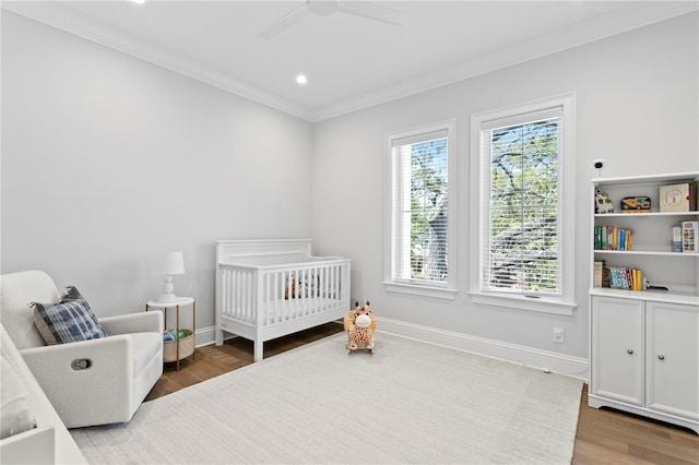 bedroom with a crib, wood finished floors, crown molding, baseboards, and ceiling fan