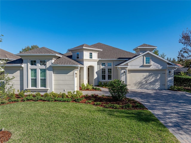 prairie-style home featuring roof with shingles, stucco siding, a front lawn, a garage, and decorative driveway