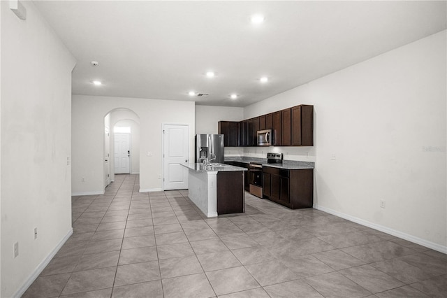 kitchen featuring stainless steel appliances, arched walkways, recessed lighting, dark brown cabinetry, and an island with sink