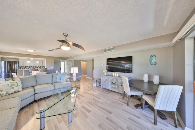 living area featuring a ceiling fan, baseboards, visible vents, light wood finished floors, and a textured ceiling