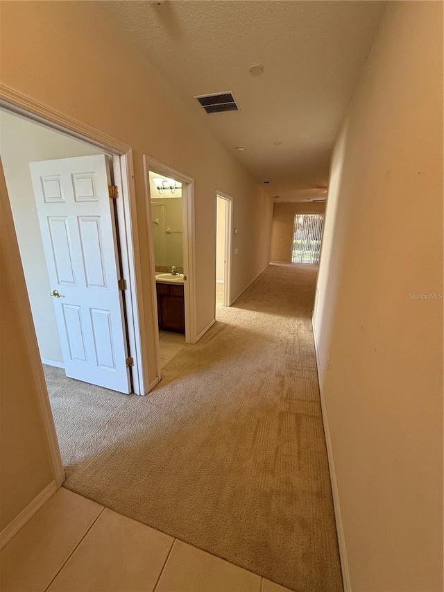 hallway with visible vents, a sink, a textured ceiling, baseboards, and light colored carpet