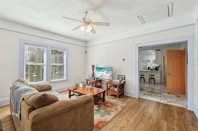 living area featuring light wood-type flooring, a textured ceiling, baseboards, attic access, and ceiling fan