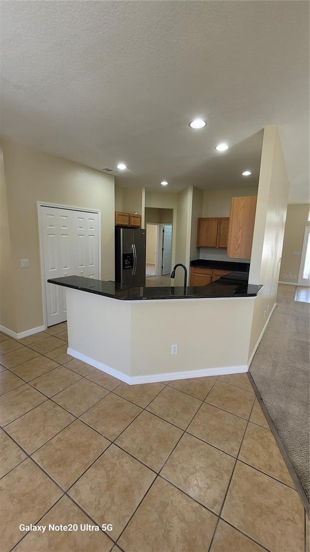 kitchen featuring refrigerator with ice dispenser, dark countertops, a peninsula, light tile patterned flooring, and brown cabinetry