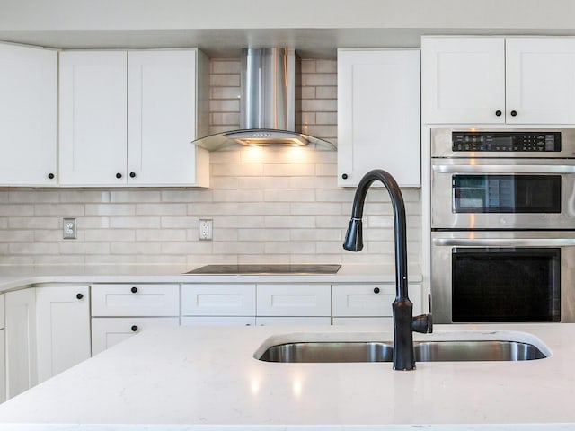 kitchen with tasteful backsplash, black electric stovetop, wall chimney range hood, stainless steel double oven, and white cabinets