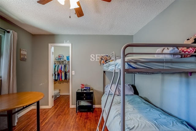 bedroom with a walk in closet, a ceiling fan, a textured ceiling, wood finished floors, and baseboards