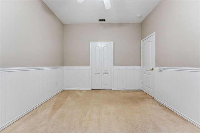 carpeted empty room with a ceiling fan, visible vents, and wainscoting