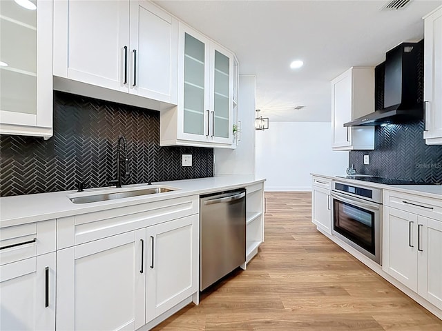 kitchen with light wood finished floors, wall chimney range hood, appliances with stainless steel finishes, white cabinetry, and a sink