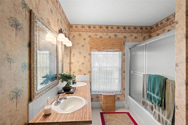 full bath with a sink, a textured ceiling, wallpapered walls, double vanity, and bath / shower combo with glass door