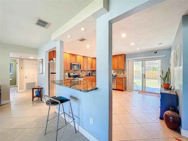 kitchen featuring visible vents, a kitchen breakfast bar, dark stone counters, appliances with stainless steel finishes, and brown cabinetry