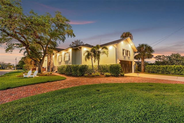 property exterior at dusk with an attached garage, a yard, driveway, and stucco siding