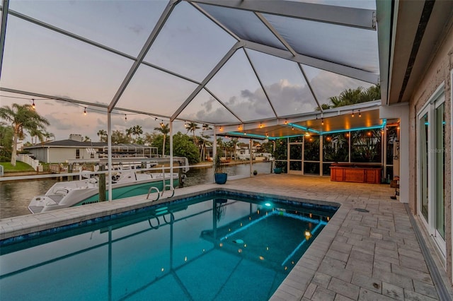 pool at dusk featuring a hot tub, a water view, a lanai, an outdoor pool, and a patio