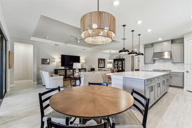 dining room with light wood-style flooring, ceiling fan with notable chandelier, a tray ceiling, recessed lighting, and baseboards