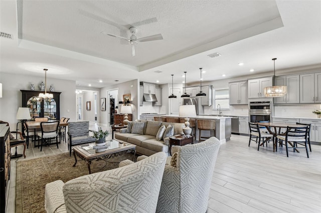 living area with visible vents, light wood finished floors, a tray ceiling, a textured ceiling, and ceiling fan with notable chandelier