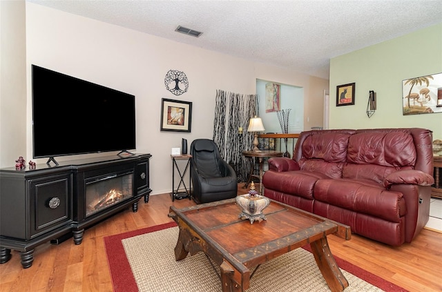 living area with visible vents, a textured ceiling, light wood-style floors, and a glass covered fireplace