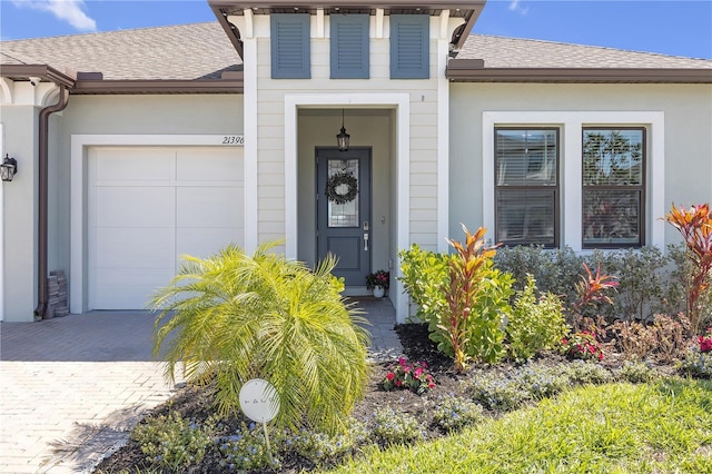 doorway to property featuring decorative driveway, roof with shingles, an attached garage, and stucco siding