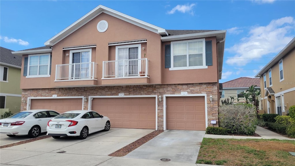 multi unit property featuring stucco siding, driveway, stone siding, an attached garage, and a balcony