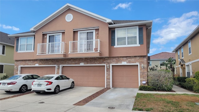 multi unit property featuring stucco siding, driveway, stone siding, an attached garage, and a balcony