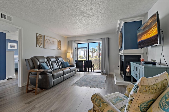 living room featuring visible vents, baseboards, a warm lit fireplace, wood finished floors, and a textured ceiling