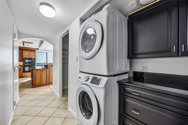 washroom featuring light tile patterned floors, stacked washing maching and dryer, arched walkways, cabinet space, and a textured ceiling