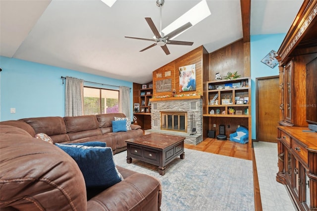 living area featuring high vaulted ceiling, a ceiling fan, wooden walls, a stone fireplace, and a skylight