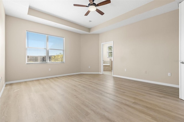 empty room featuring a raised ceiling, light wood-style flooring, a ceiling fan, and baseboards