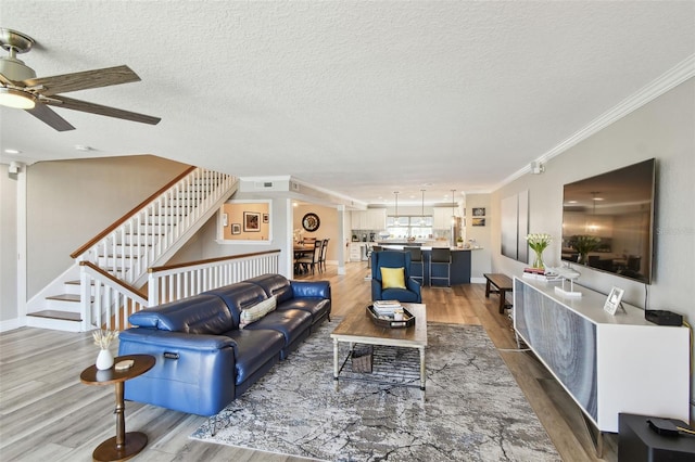 living area with a ceiling fan, a textured ceiling, wood finished floors, crown molding, and stairs