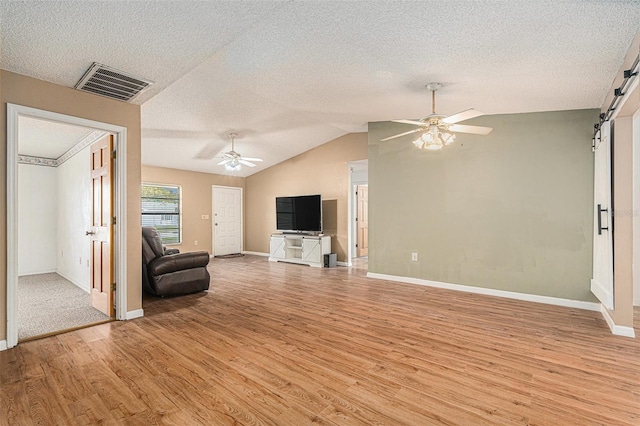 unfurnished living room with visible vents, ceiling fan, light wood-type flooring, a barn door, and vaulted ceiling