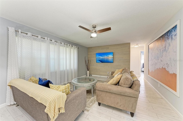 living area featuring a ceiling fan, a textured ceiling, light wood finished floors, baseboards, and an accent wall