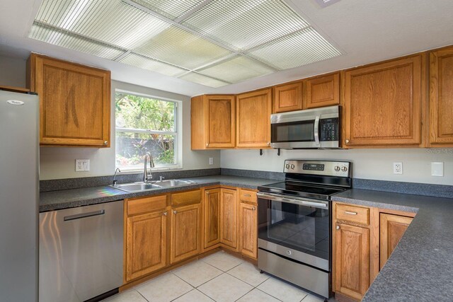 kitchen with light tile patterned floors, brown cabinetry, a sink, stainless steel appliances, and dark countertops