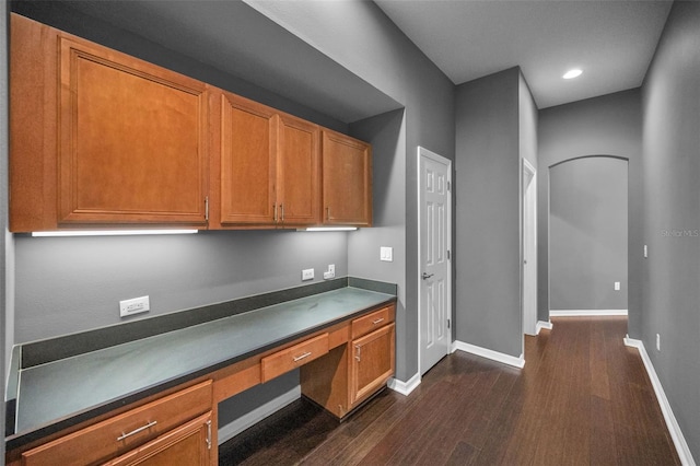 kitchen featuring brown cabinetry, baseboards, dark wood finished floors, built in desk, and dark countertops