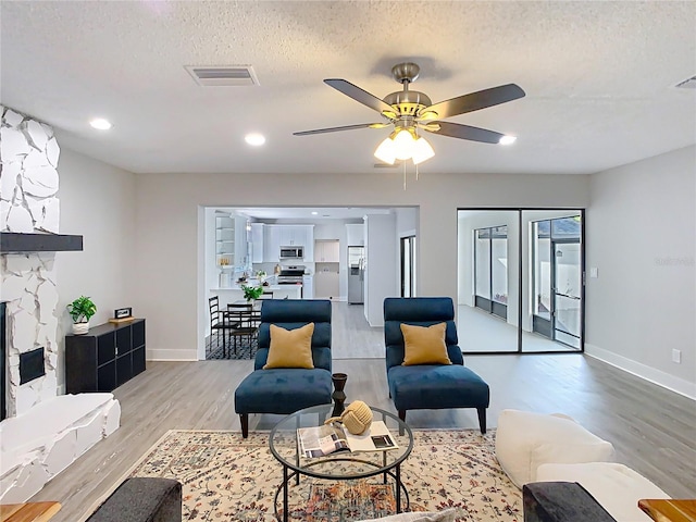 living area featuring light wood-type flooring, visible vents, a textured ceiling, a stone fireplace, and ceiling fan