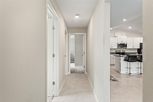 hallway with light tile patterned floors, baseboards, visible vents, vaulted ceiling, and a textured wall