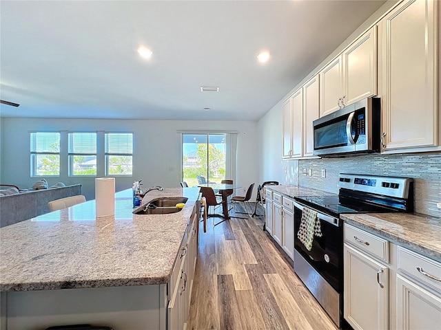 kitchen featuring light wood finished floors, an island with sink, a sink, decorative backsplash, and stainless steel appliances