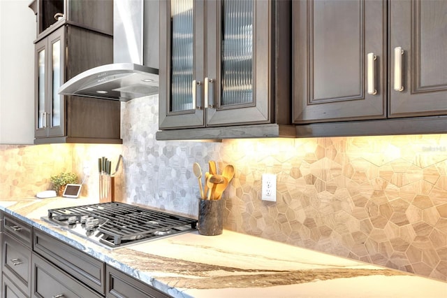 kitchen featuring stainless steel gas stovetop, decorative backsplash, glass insert cabinets, wall chimney exhaust hood, and light stone counters