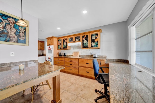 kitchen with under cabinet range hood, glass insert cabinets, light stone countertops, and backsplash