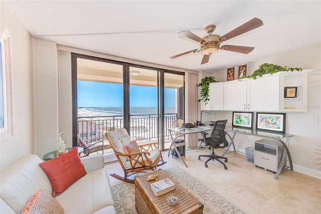 office area with a water view, a ceiling fan, expansive windows, a view of the beach, and baseboards