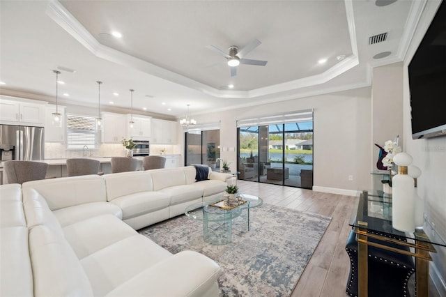 living area with visible vents, ceiling fan with notable chandelier, a tray ceiling, crown molding, and light wood finished floors