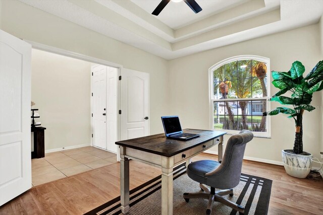 office featuring baseboards, a tray ceiling, ceiling fan, and light wood-type flooring
