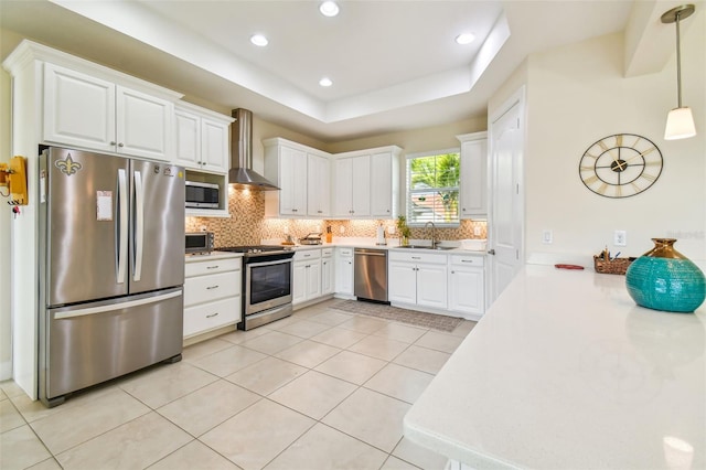 kitchen with tasteful backsplash, wall chimney range hood, appliances with stainless steel finishes, white cabinets, and a raised ceiling