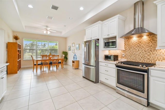 kitchen with visible vents, wall chimney range hood, a tray ceiling, appliances with stainless steel finishes, and a ceiling fan