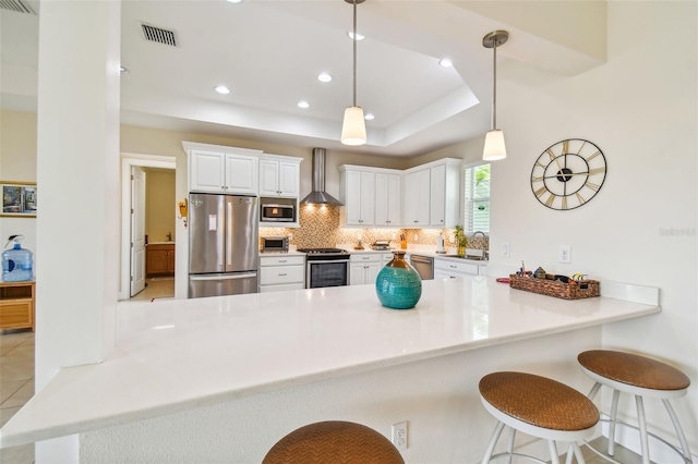 kitchen featuring a peninsula, stainless steel appliances, wall chimney exhaust hood, and a raised ceiling