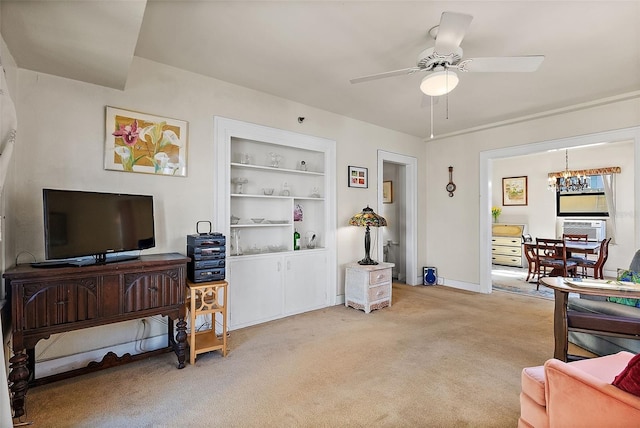 living area featuring built in shelves, ceiling fan with notable chandelier, baseboards, and light carpet