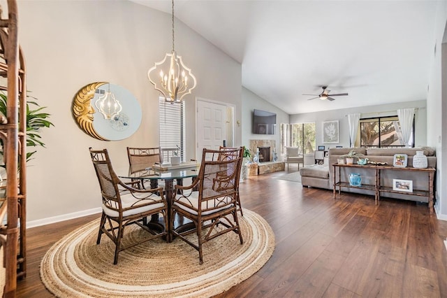 dining space featuring baseboards, a stone fireplace, ceiling fan with notable chandelier, dark wood-style floors, and high vaulted ceiling