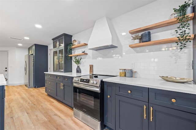 kitchen featuring light wood-type flooring, custom range hood, decorative backsplash, appliances with stainless steel finishes, and open shelves